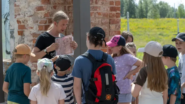 Pikkade juustega mees näitab laste grupile ajalooliste varemete juures vana kaarti ja selgitusi.